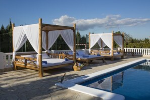 Un espace aménagé au bord de la piscine, avec des chaises longues en bois et des auvents blancs, une terrasse dallée de pierre et une piscine à l’eau d’un bleu cristallin.