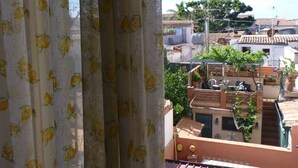 Vista a través de una ventana con cortinas florales, que muestra una terraza en la azotea con plantas en macetas y un tejado de baldosas.