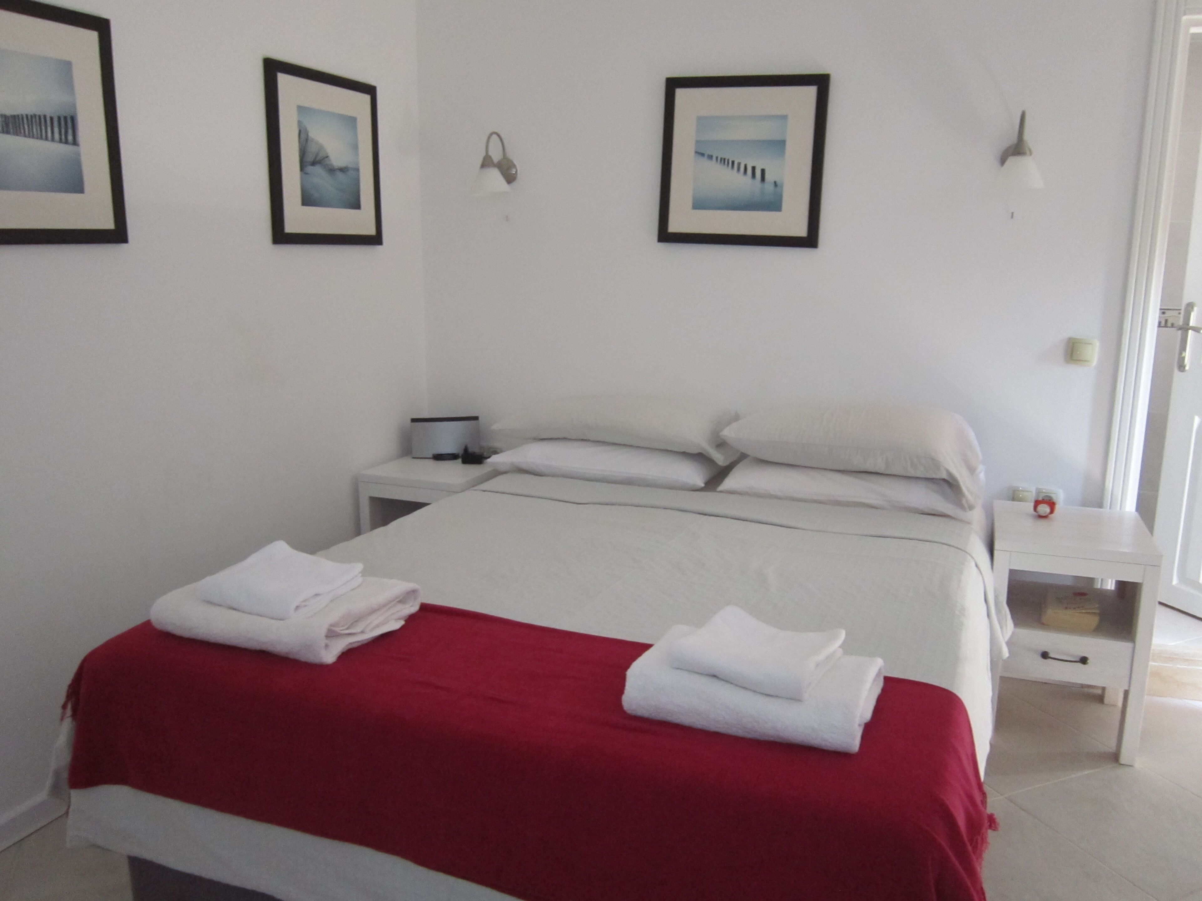 A neatly made bed with a red blanket, white pillows, and two folded towels. There are two bedside tables, one with a lamp and the other with a small clock. The room has white walls and three framed pictures on the wall.
