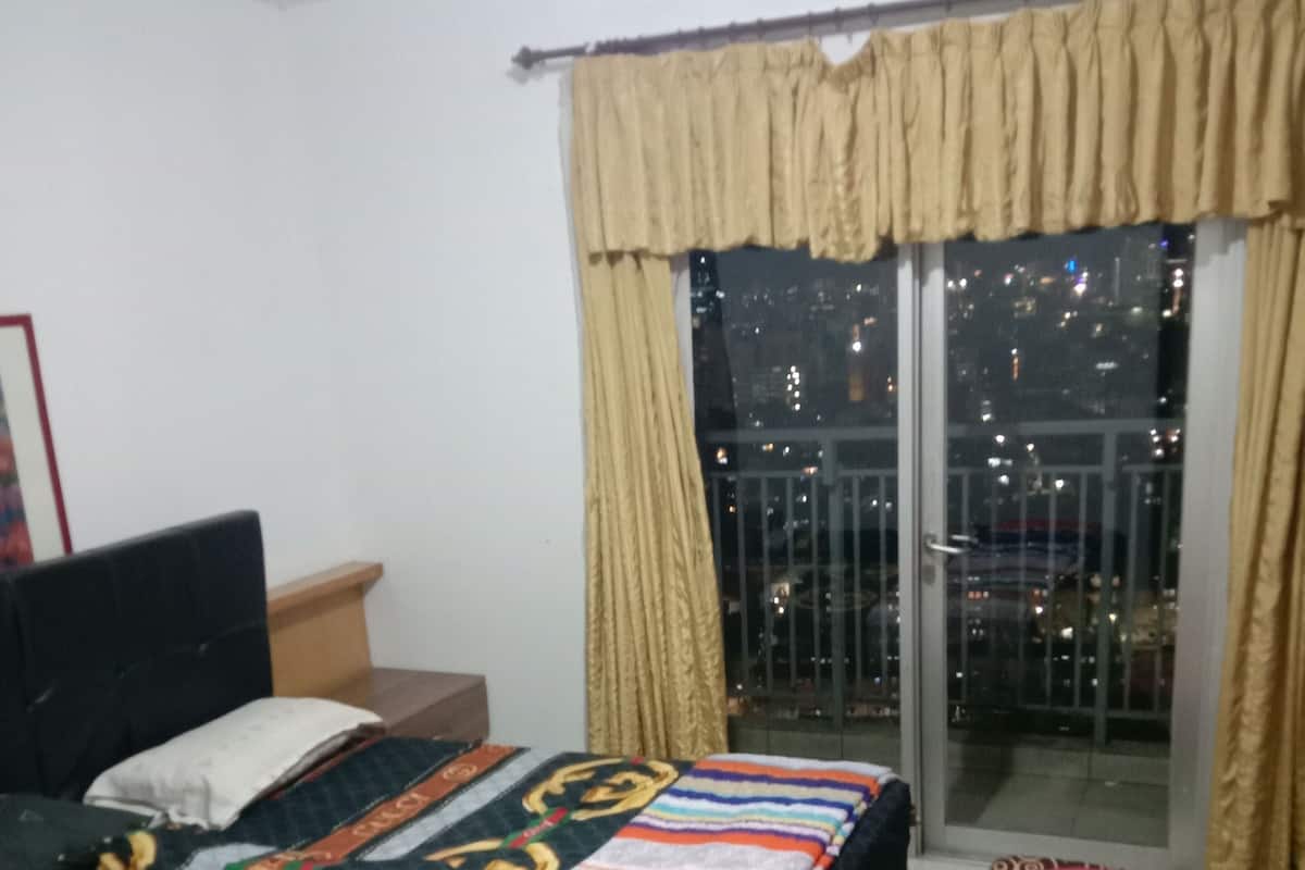 A bedroom with a bed, a nightstand, a window with curtains, and a view of the city at night.