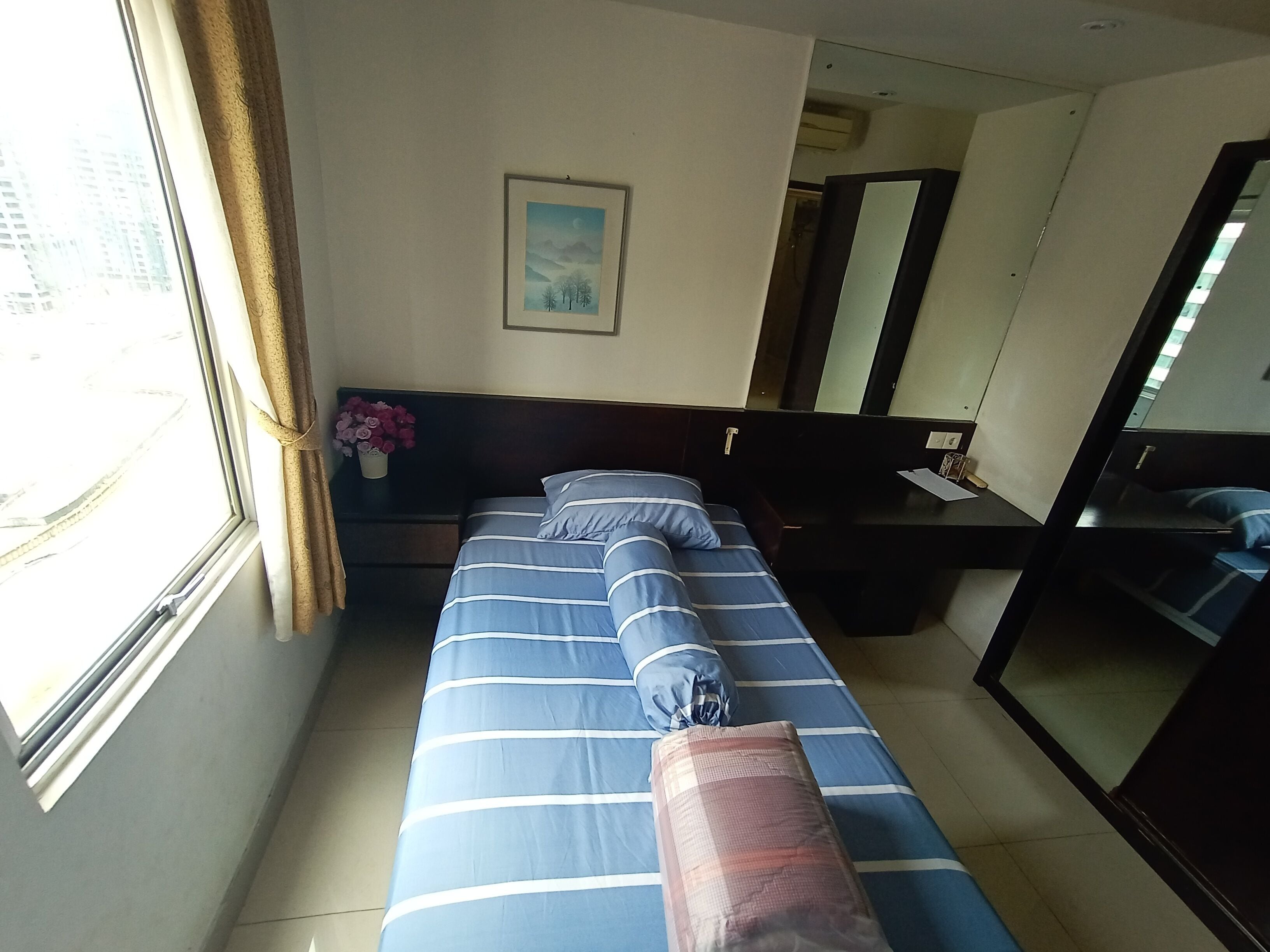 A hotel room with a bed, a bedside table, a mirror, a window with curtains, and a painting on the wall.