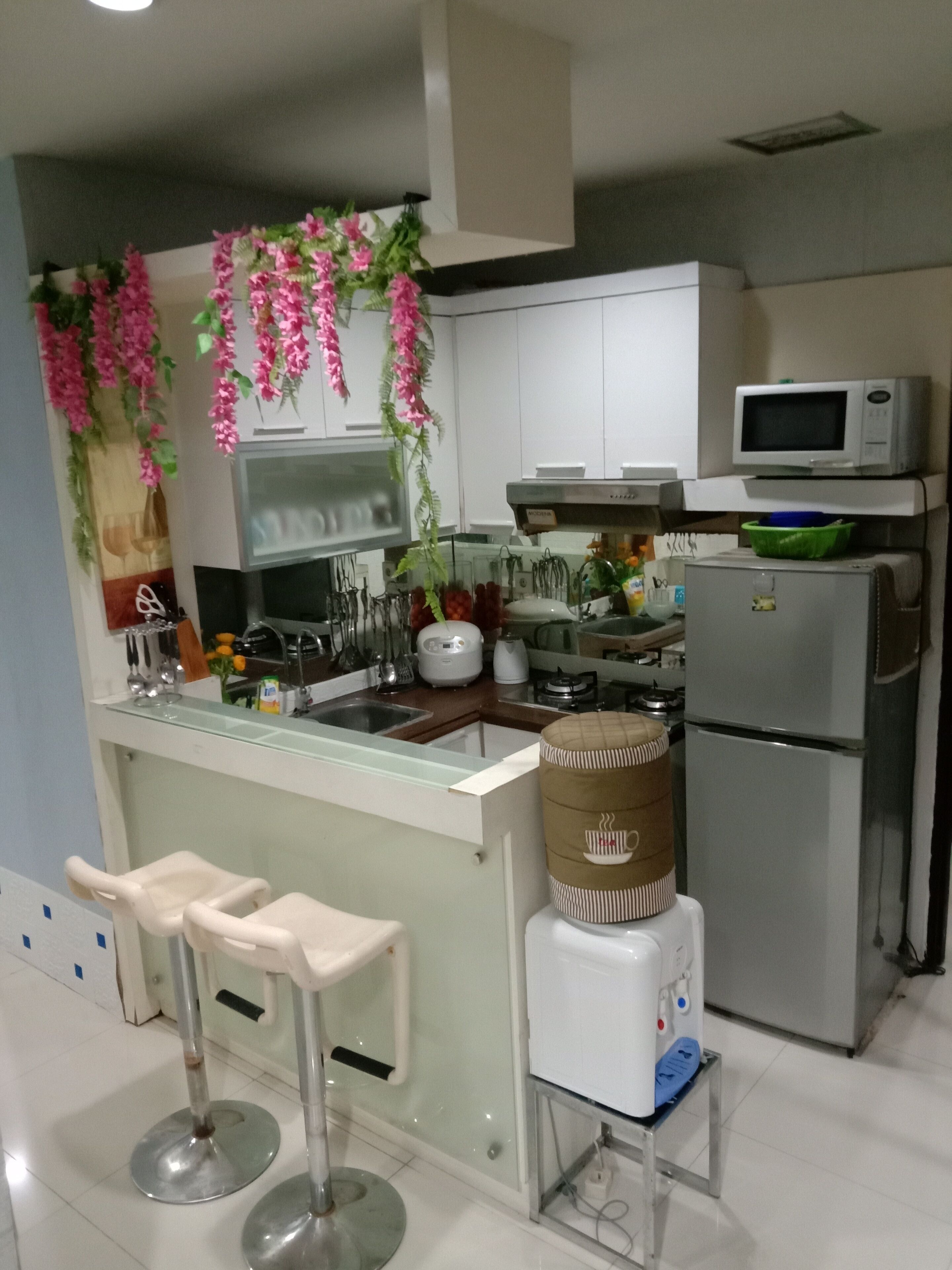 A modern kitchen with white cabinets, a stainless steel refrigerator, a microwave, and a sink. There are two bar stools and a decorative plant hanging from the ceiling.