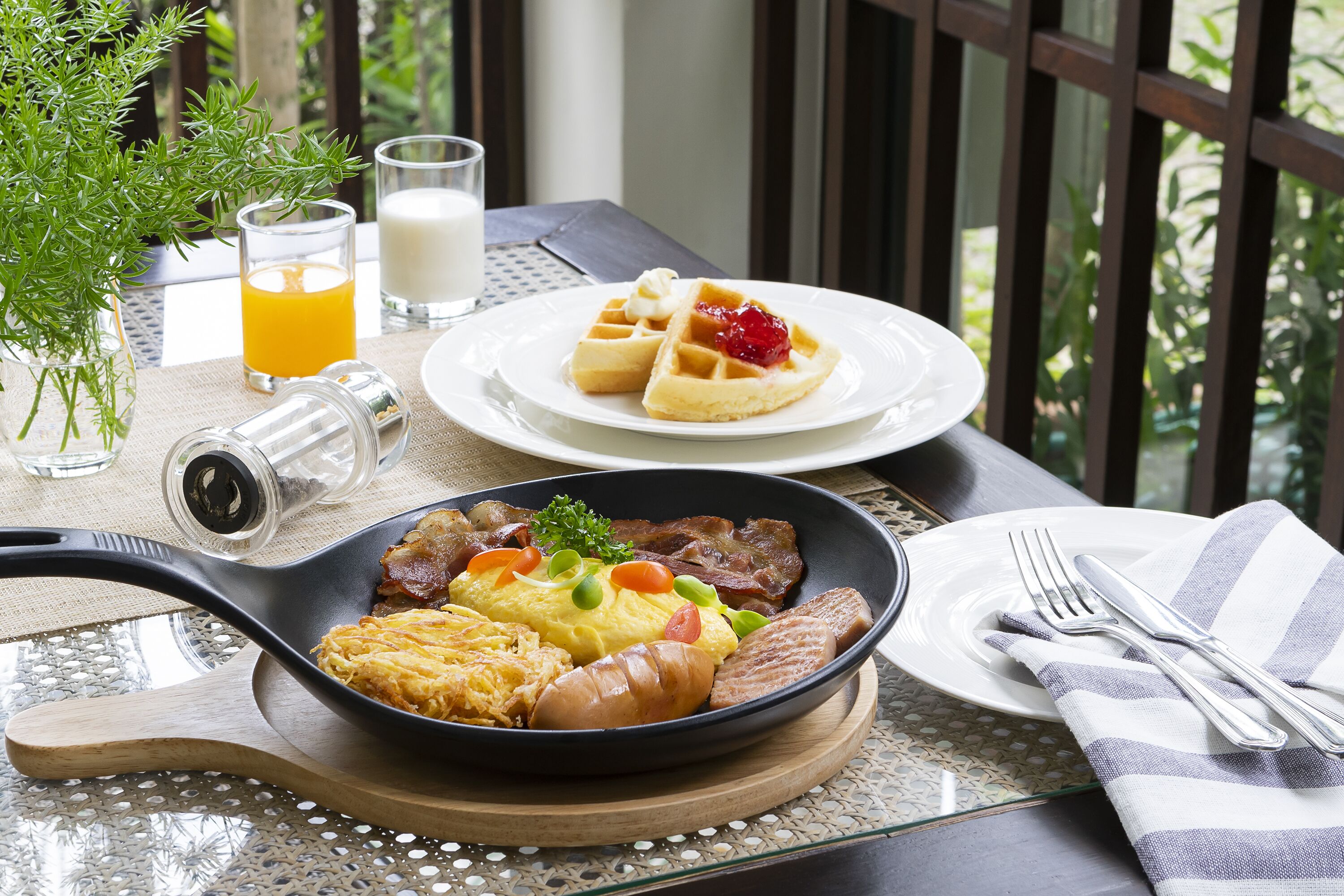 A breakfast setting with a plate of waffles, eggs, and bacon, a bowl of scrambled eggs, and a glass of orange juice on a table with a view of greenery.