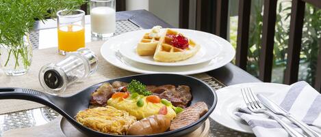 A breakfast setting with a plate of waffles, eggs, and bacon, a bowl of scrambled eggs, and a glass of orange juice on a table with a view of greenery.