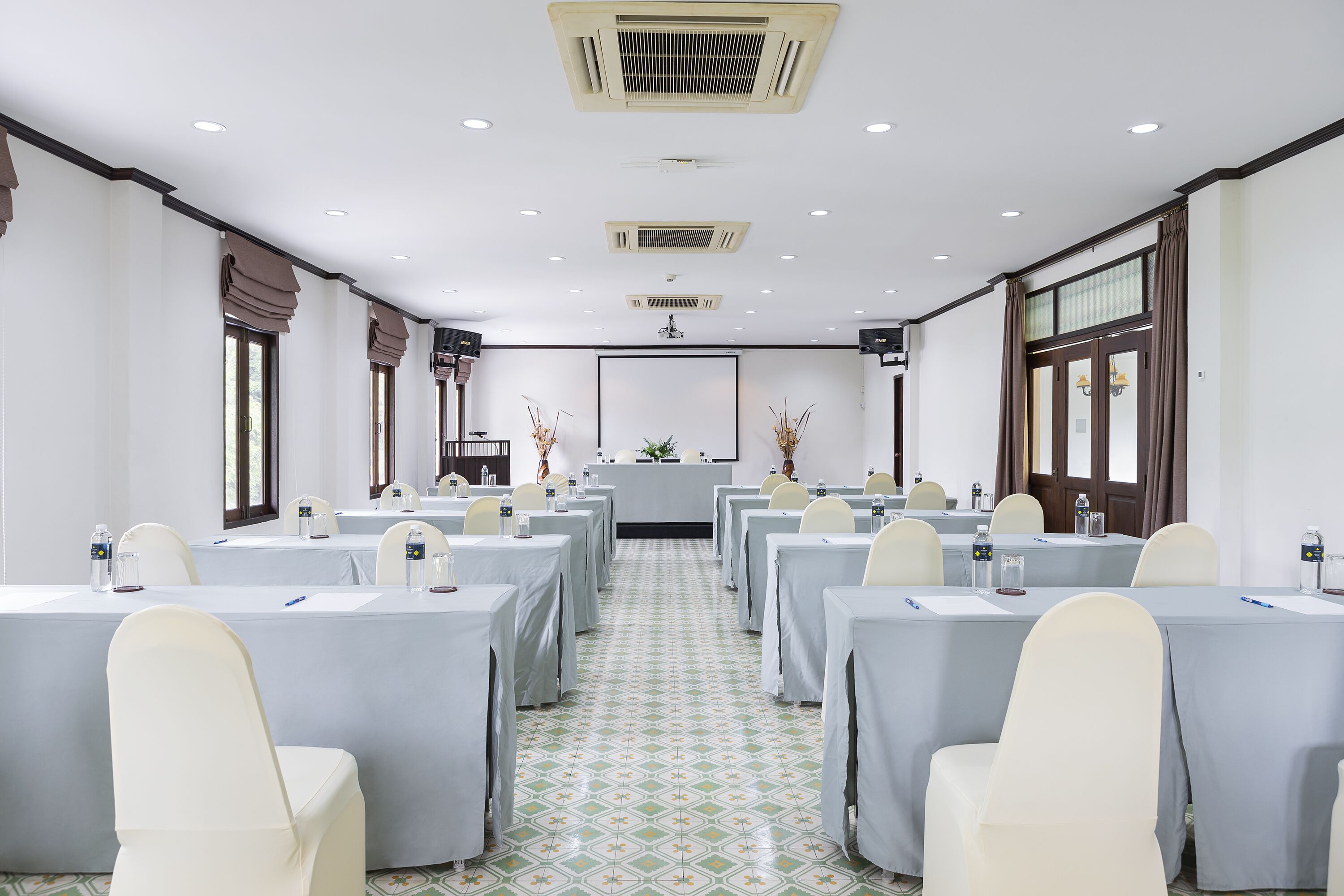A conference room with multiple tables and chairs, a central podium, and large windows with curtains.