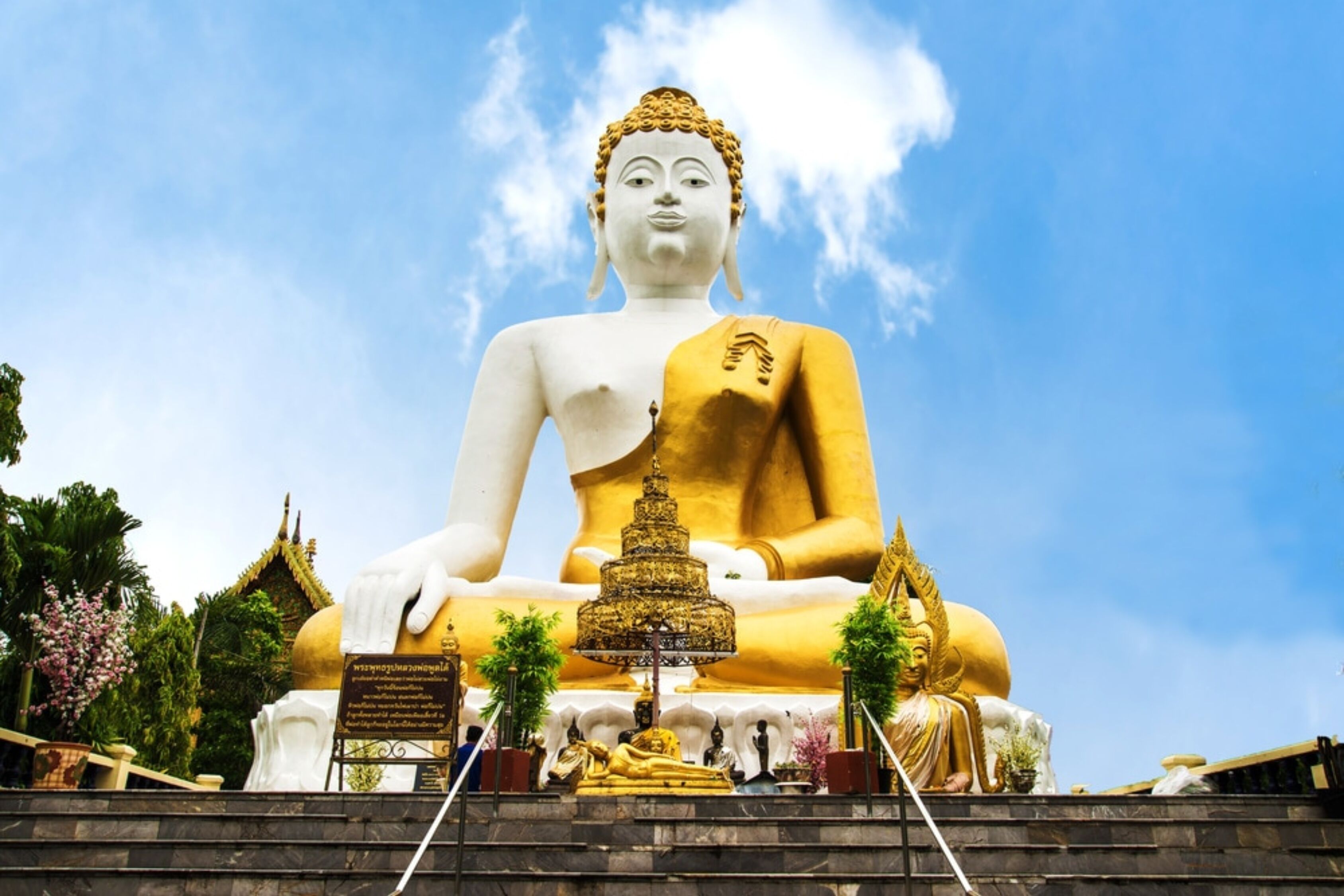 Una gran estatua dorada de Buda con una estupa dorada en primer plano, rodeada de estatuas más pequeñas y una estructura tradicional de tejado tailandés.