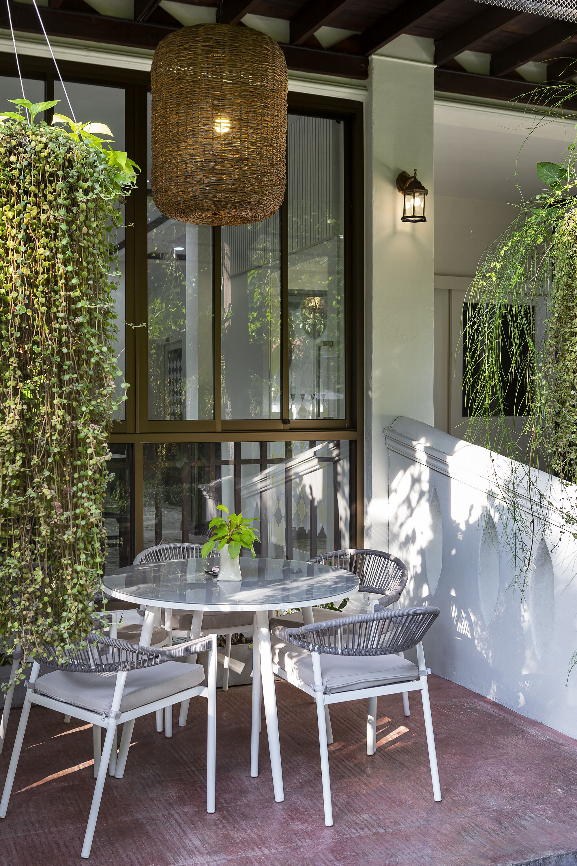 A round glass table with white chairs, a hanging plant, and a wicker pendant light in a modern outdoor setting.