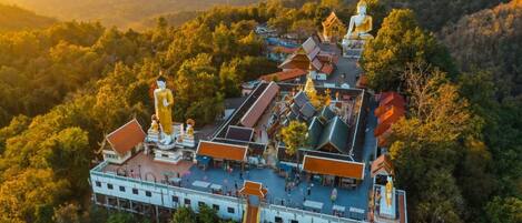 An aerial view of a temple complex with multiple buildings, including a large golden statue, surrounded by lush greenery and hills in the background.