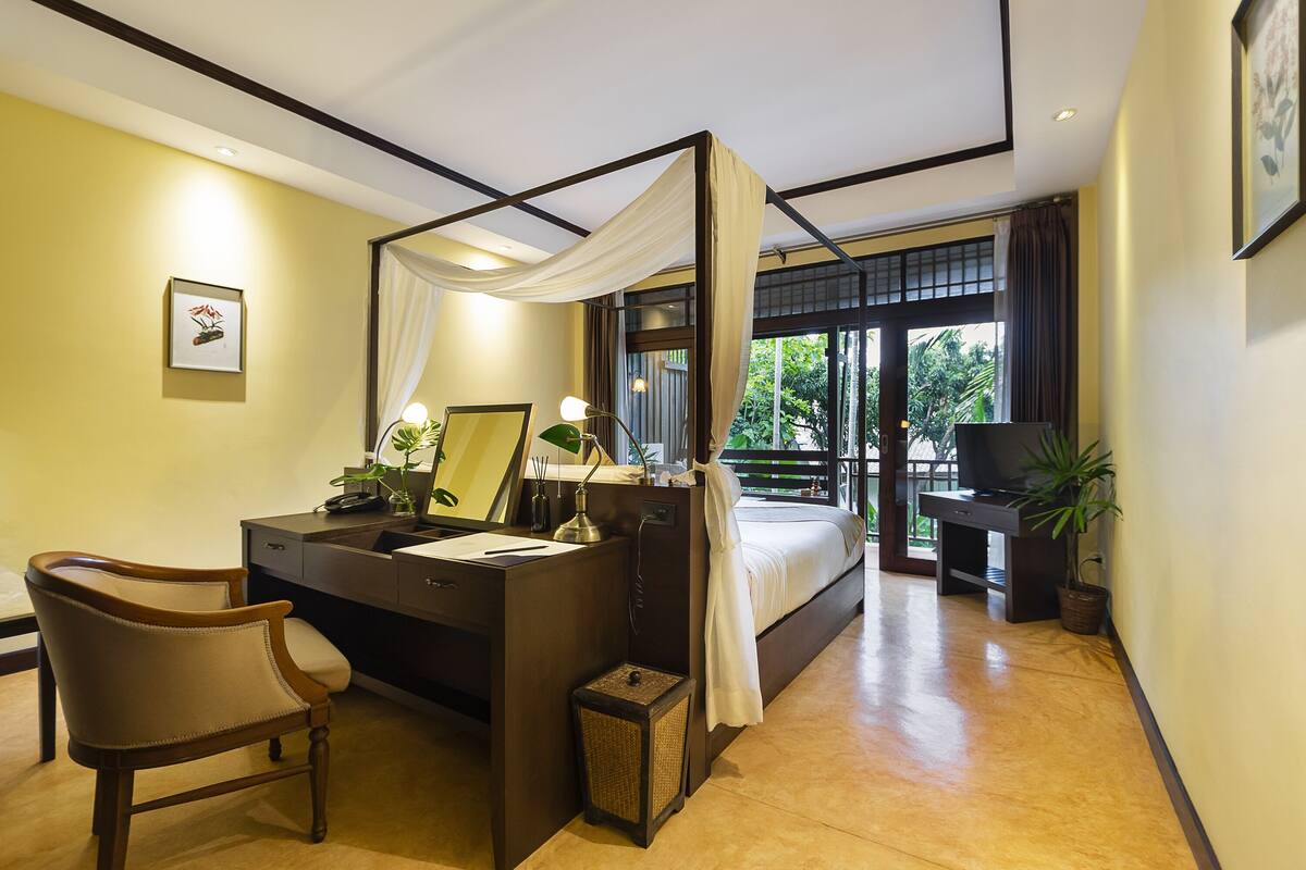 A hotel room with a bed, desk, chair, bedside table, and a potted plant.