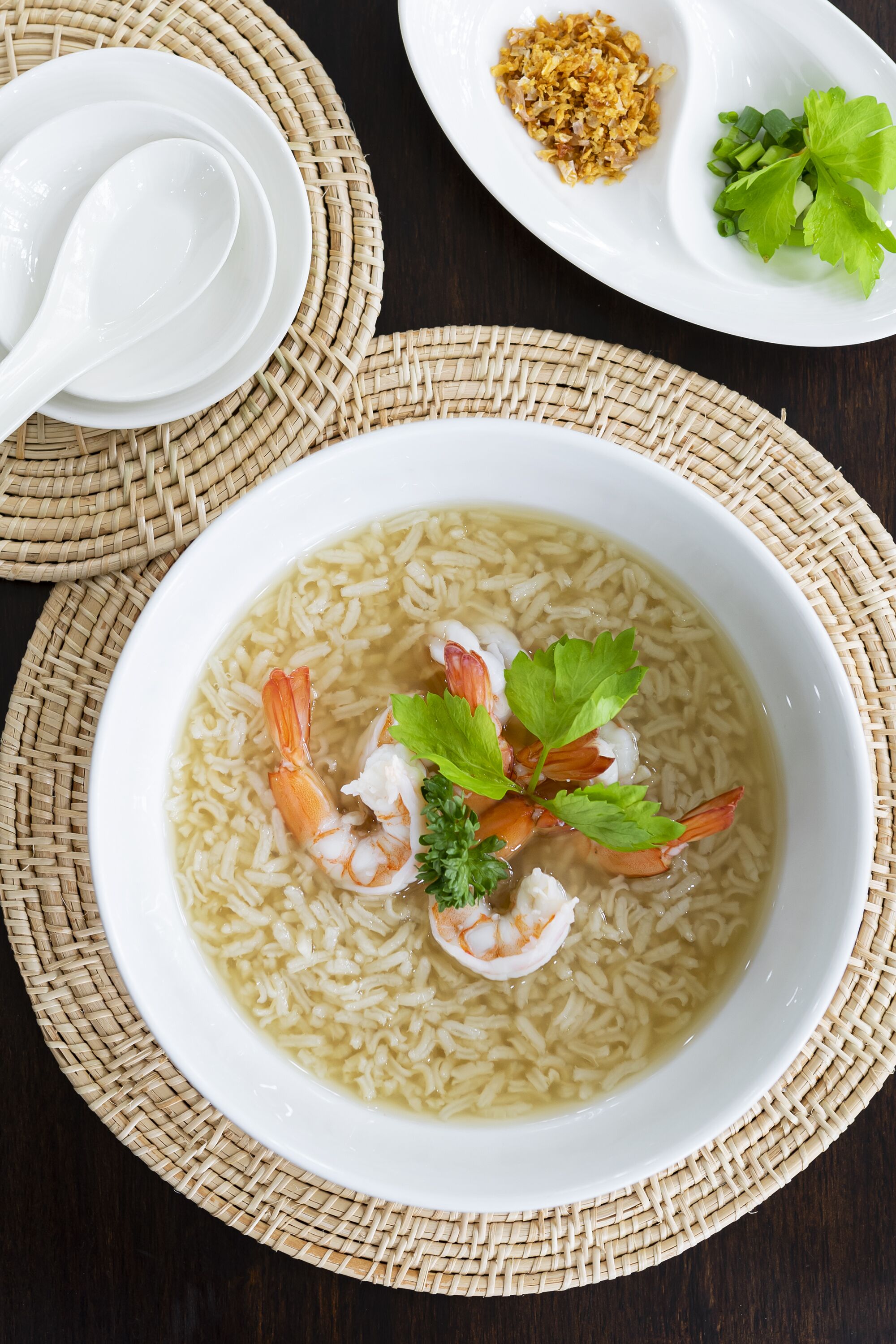Un tazón de sopa de arroz con camarones y adorno de perejil, acompañado de un pequeño plato de condimentos y una cuchara.