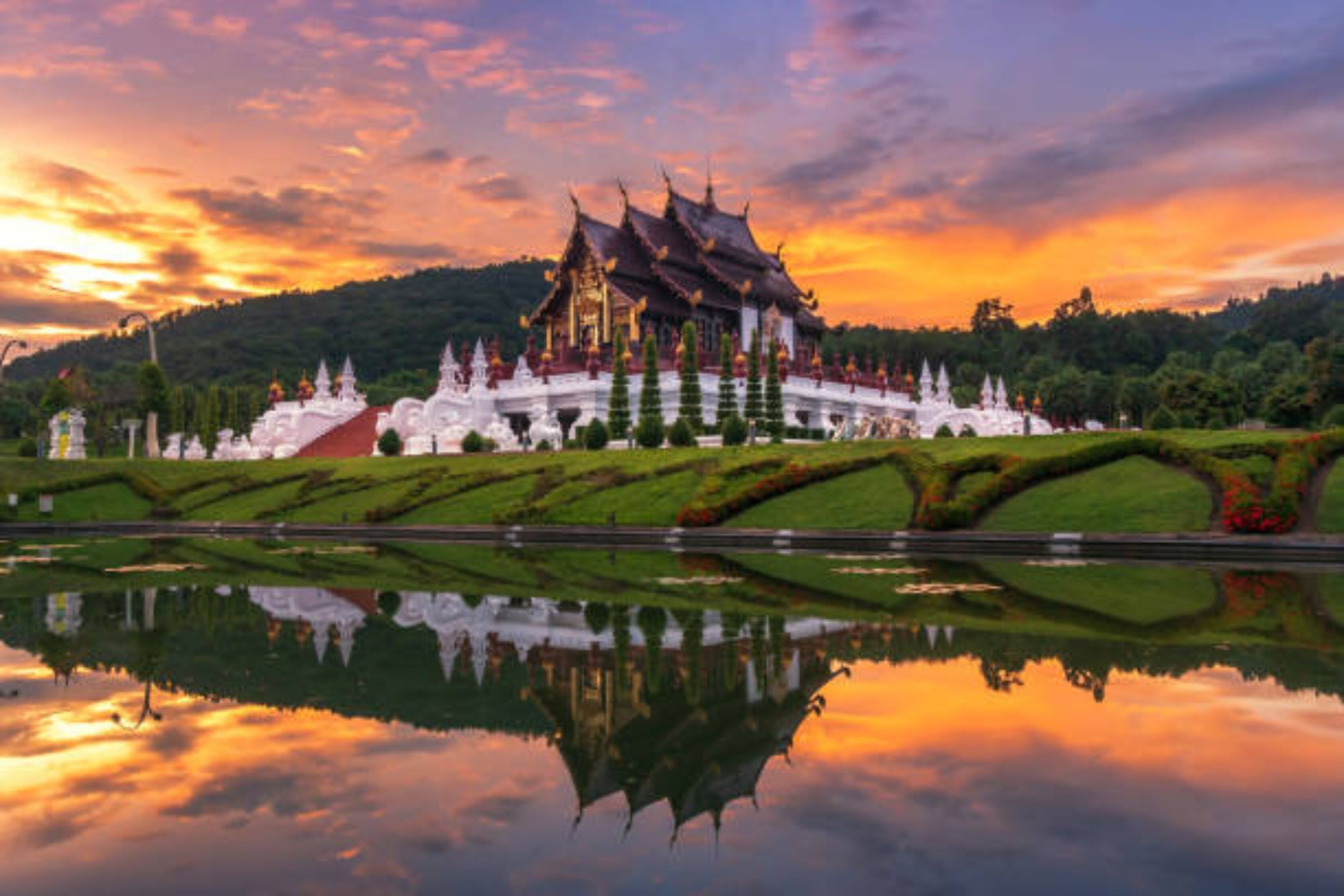 A traditional multi-tiered temple with a reflection in a calm body of water, surrounded by manicured gardens and a hilly backdrop.