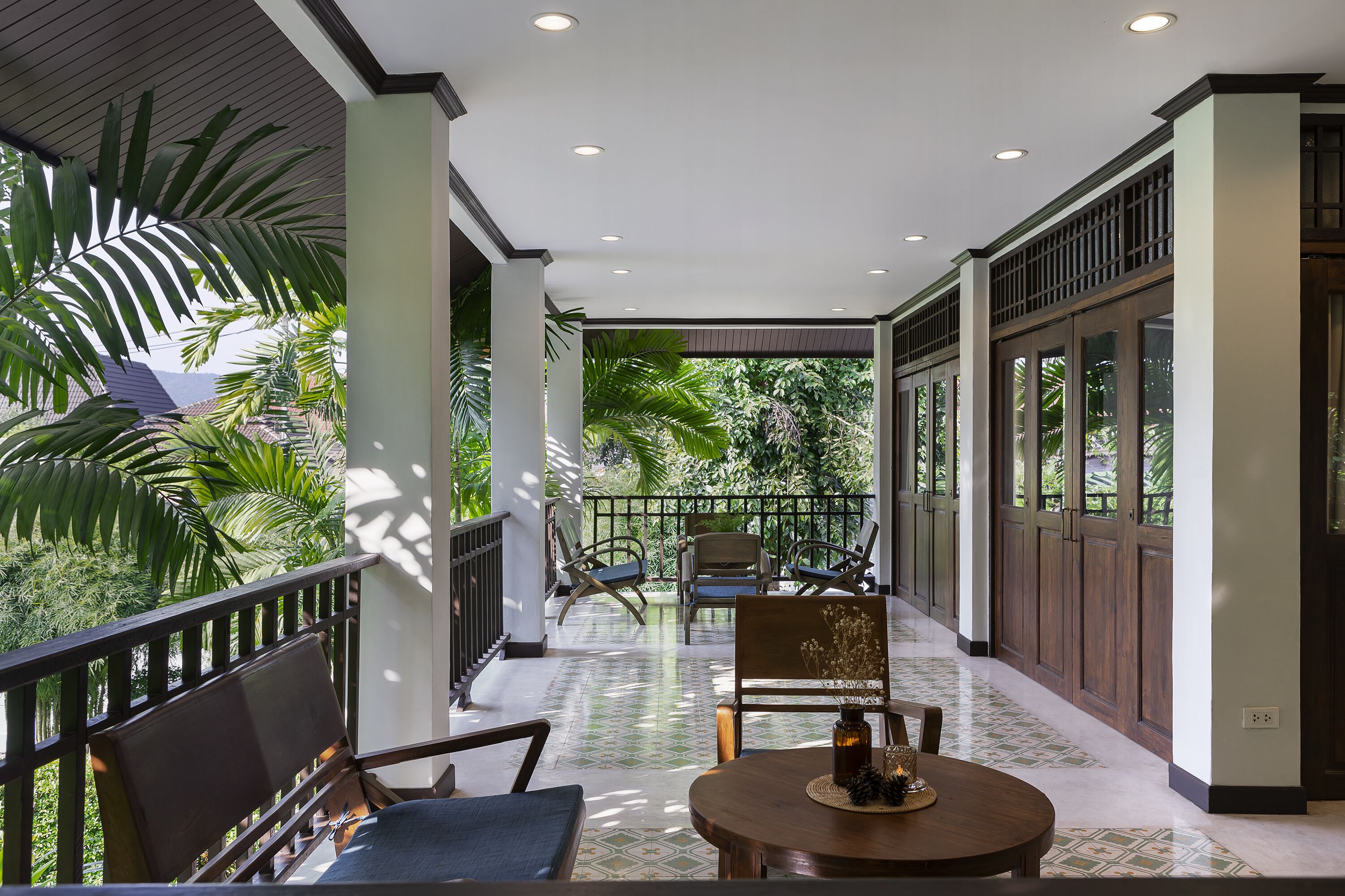 A covered outdoor seating area with a table and chairs, surrounded by large windows and doors, offering views of greenery.