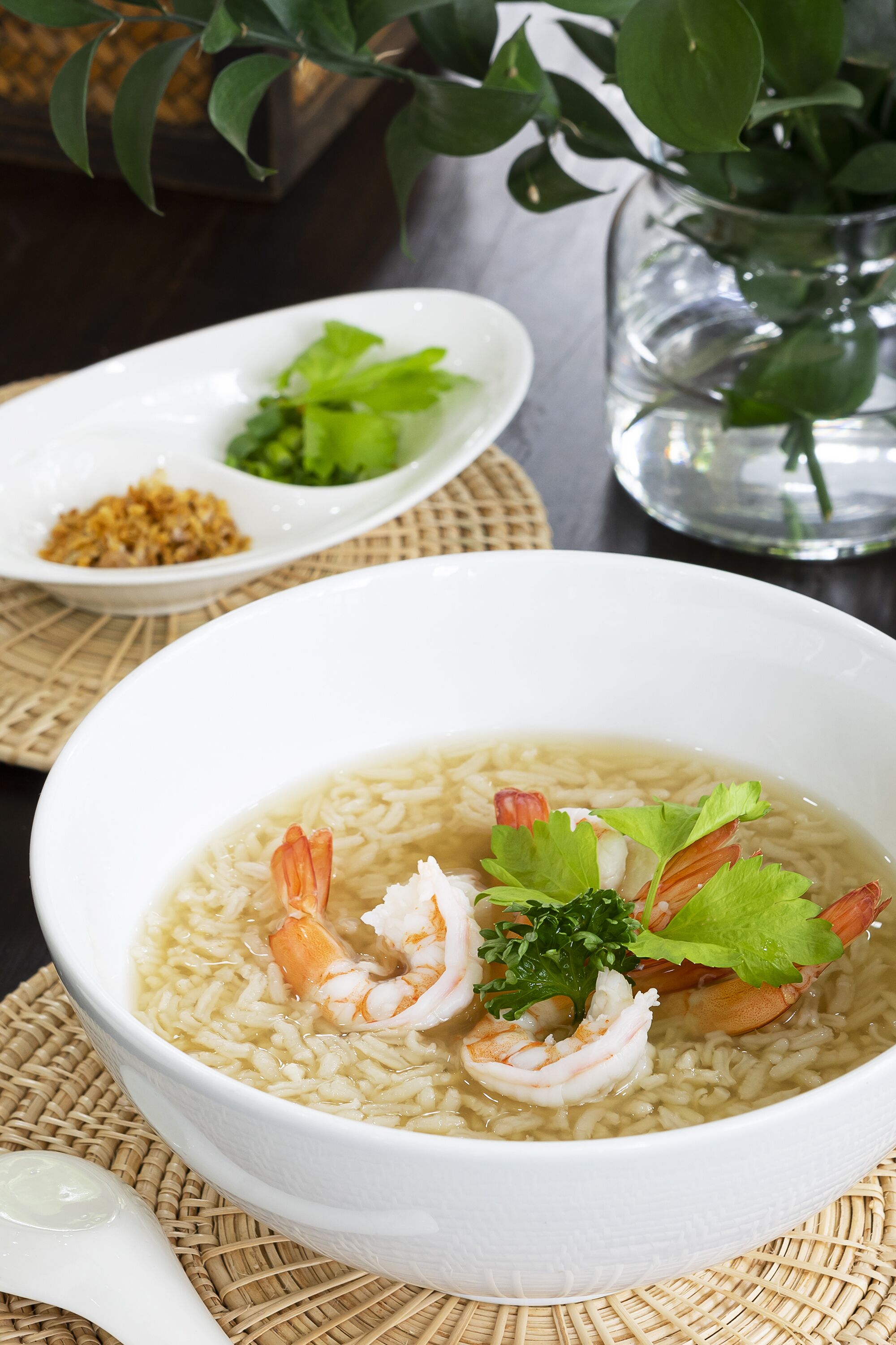 A bowl of rice with shrimp and greens, a white spoon, and a small dish with greens and spices on a woven mat.