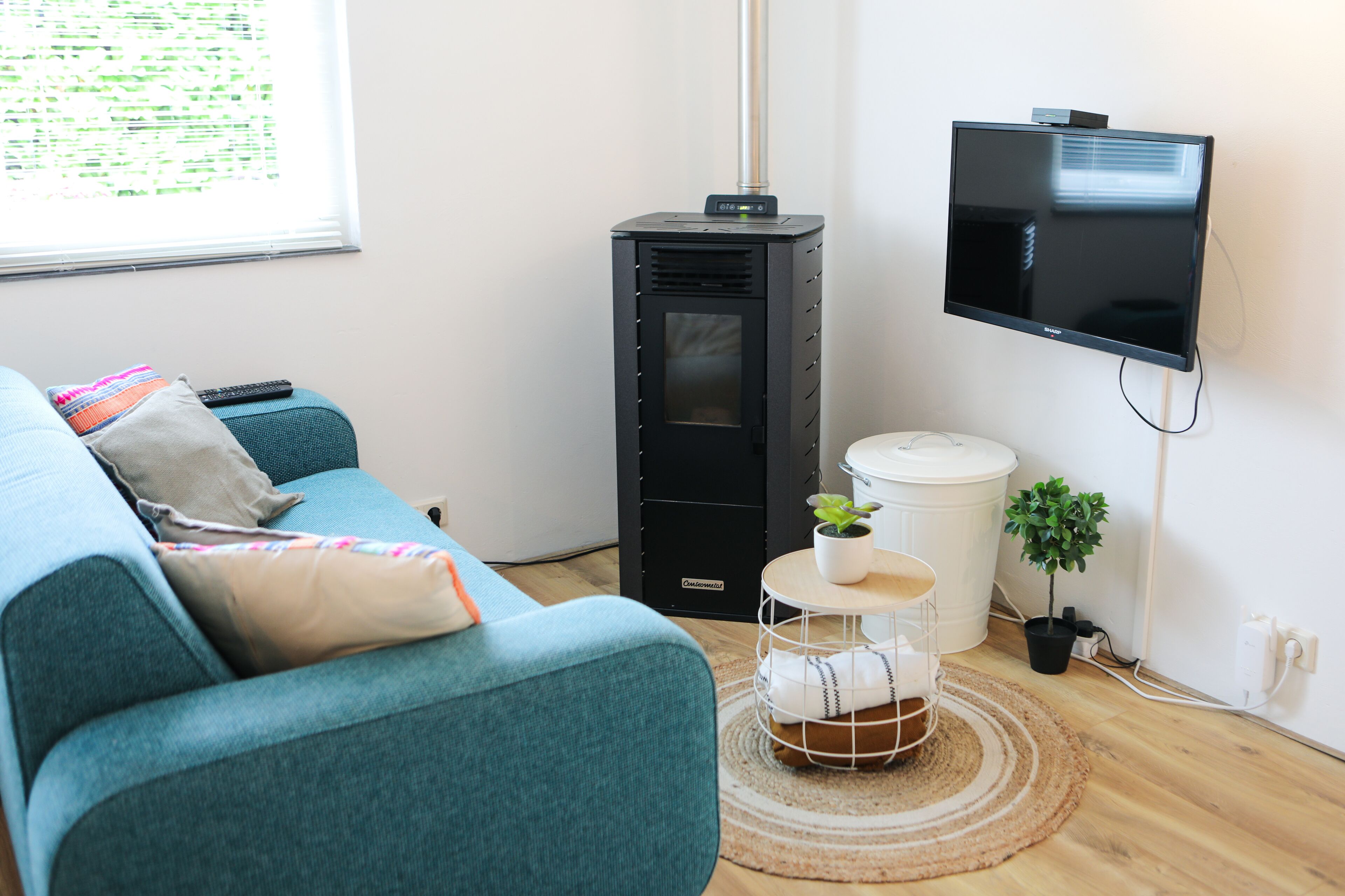 Una sala moderna con un sofá color esmeralda, un calefactor negro, un televisor montado, una mesita redonda con una planta y un cubo de basura blanco.