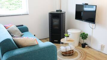 Una sala moderna con un sofá color esmeralda, un calefactor negro, un televisor montado, una mesita redonda con una planta y un cubo de basura blanco.