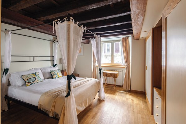 A bedroom with a four-poster bed, wooden flooring, a window with curtains, and a bedside table.