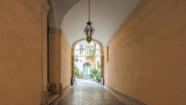 A corridor with a high ceiling, a hanging light fixture, and a view of a building through an arched doorway. - Elegant two-bedroom apartment in the very heart of Rome. Perfect for a family! (Roma)
