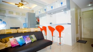 A modern living room with a black sofa, colorful cushions, and a glass partition separating the living area from the kitchen. - SM Jazz Residence by Good Rent (Makati)