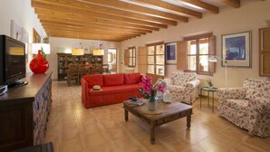 Uma sala de estar ampla, com teto de madeira, um sofá vermelho, poltronas florais e uma mesa de centro com objetos decorativos.
