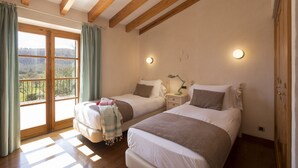 Quarto com duas camas, uma janela grande, vigas de madeira e uma mesa de cabeceira com abajur.