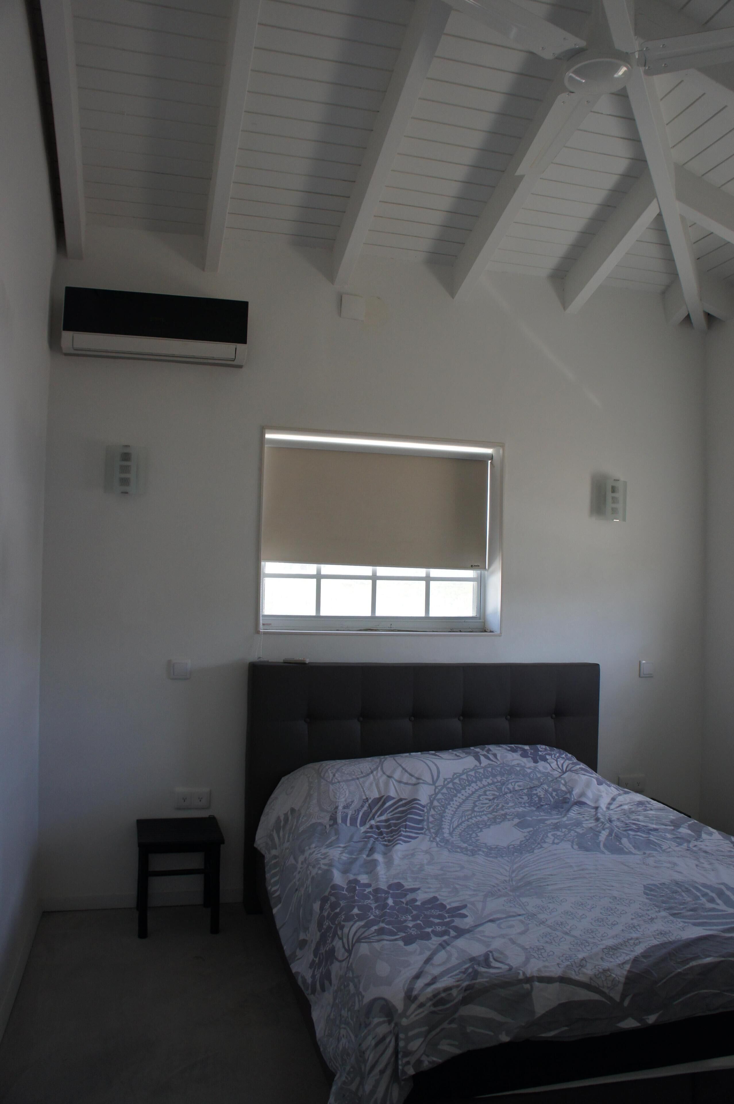 A bedroom with a bed, a small table, a window with blinds, and a ceiling fan.