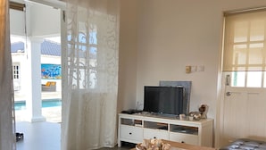 A living room with a wooden table, a television, and a sofa. There is a window with curtains and a view of the outside. - Villa Sol, luxury fully equipped villa with wifi, a short distance from the sea (Kralendijk)