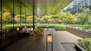 An outdoor seating area with wooden flooring, chairs, and tables, surrounded by glass walls and a view of a garden with trees and a modern building in the background. - The PuXuan Hotel and Spa (Beijing)