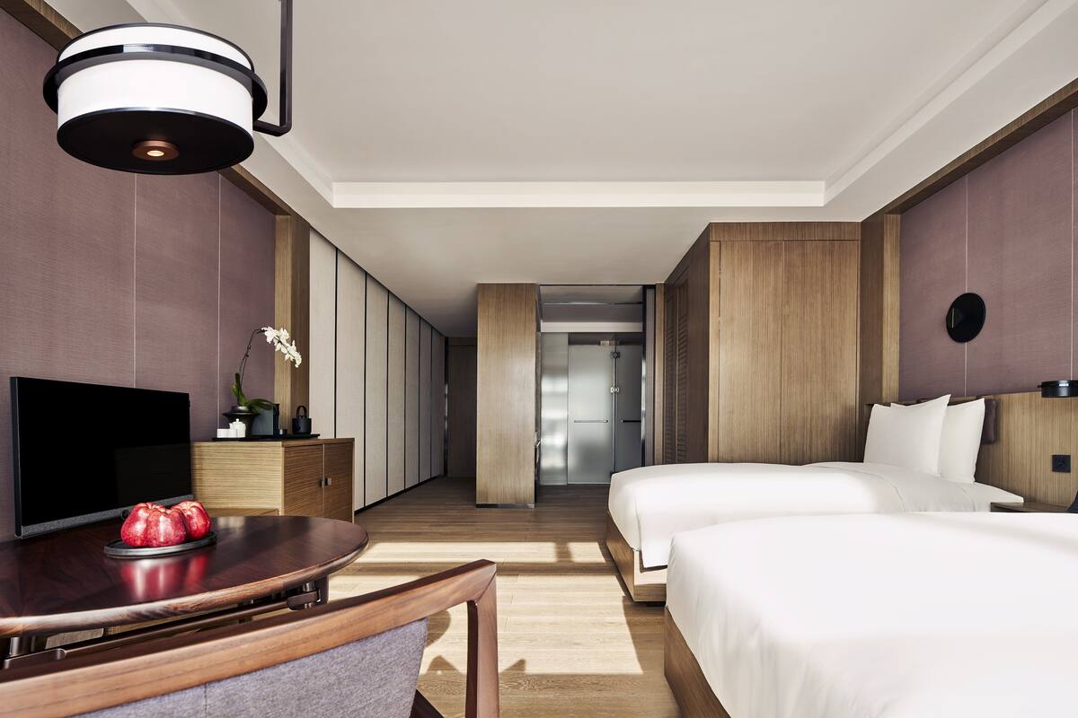 A hotel room with two beds, a flat-screen TV, a wooden desk, and a modern light fixture.