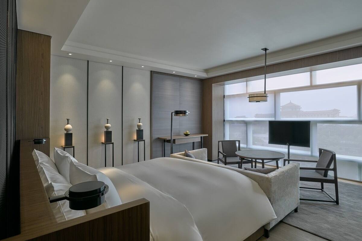 A modern hotel room with a large bed, a desk, a chair, a TV, and a view of a cityscape.