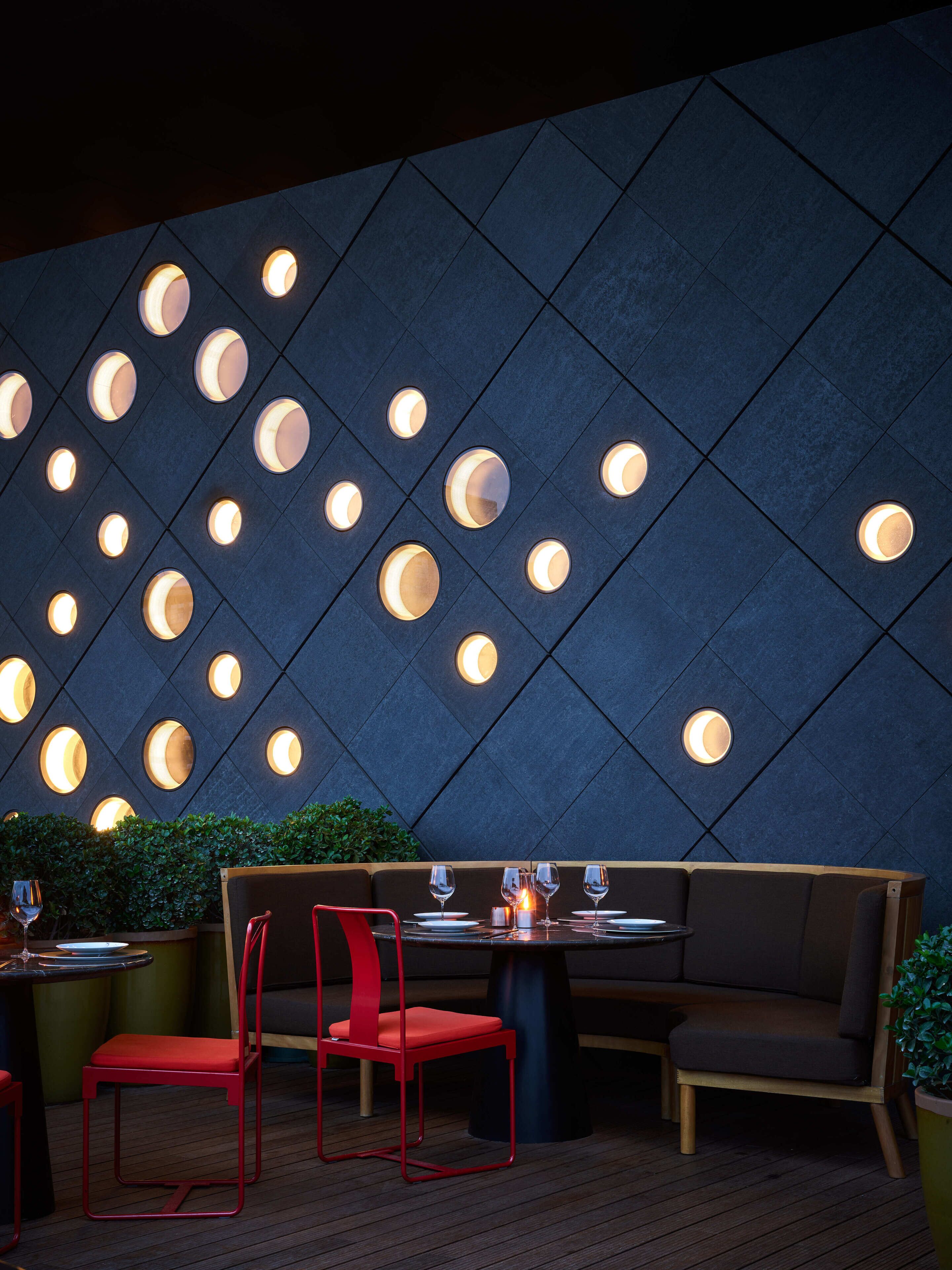 Un restaurant avec un mur sombre orné de luminaires circulaires et une table en bois entourée de chaises rouges.