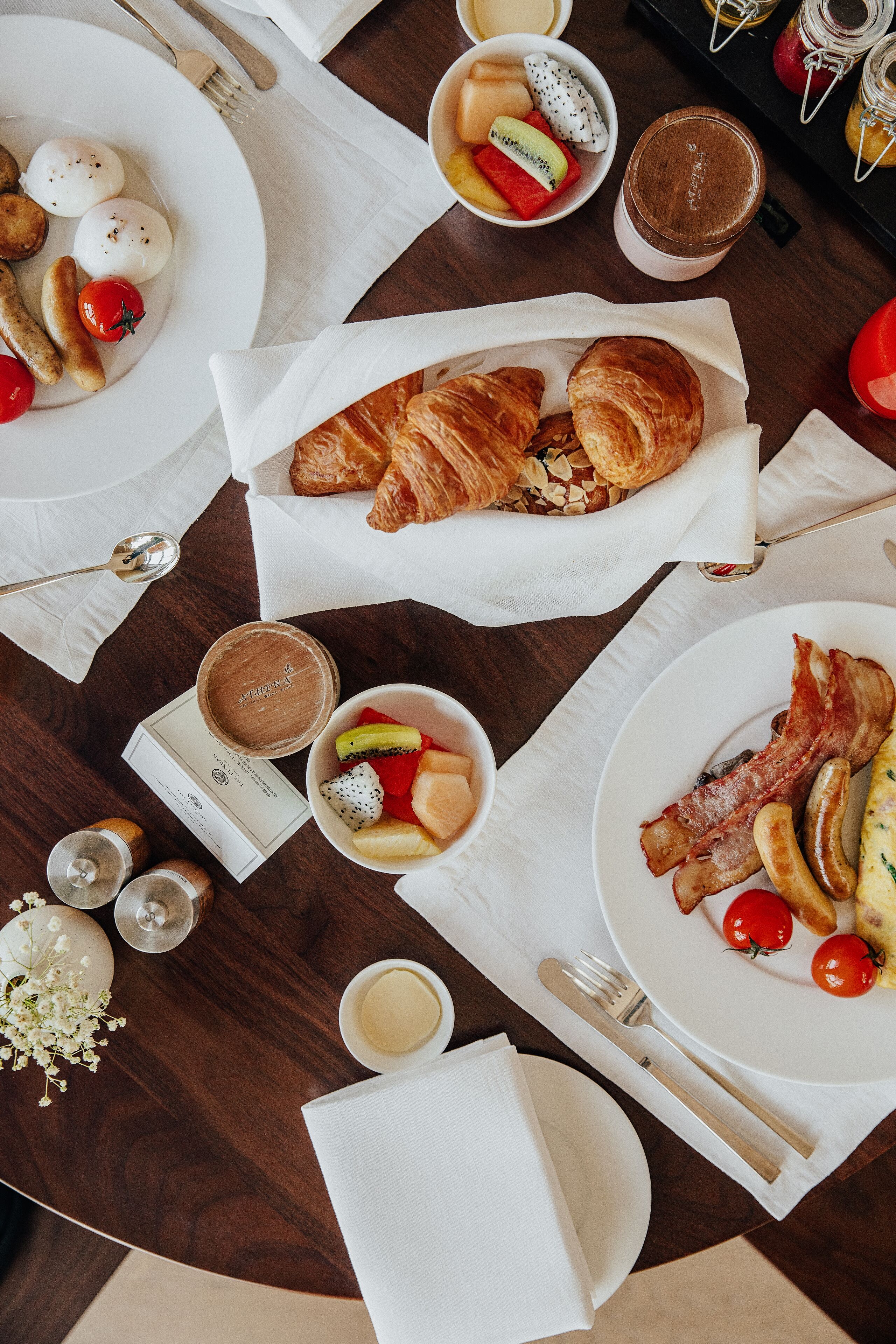 A breakfast setting with plates of food, including croissants, sausages, and tomatoes, along with a cup of coffee and a jar of jam.