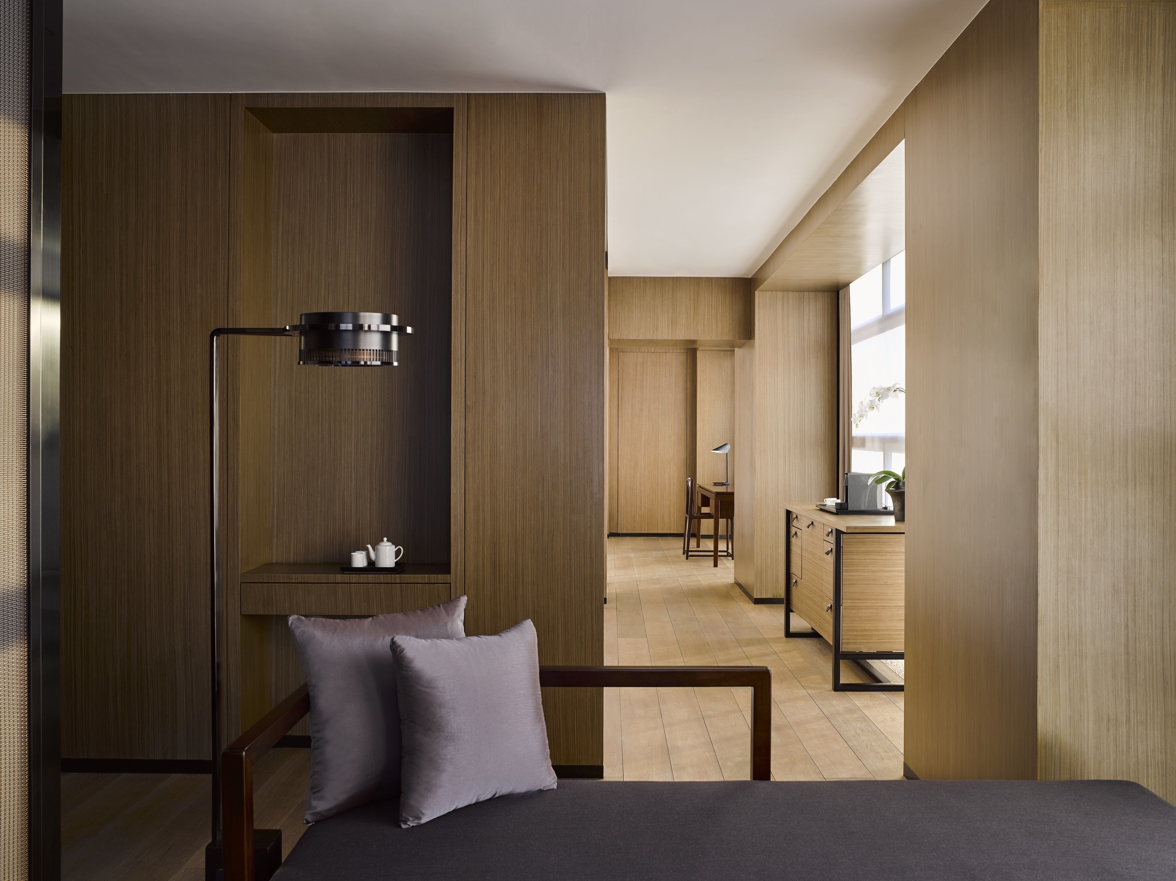 A modern hotel room with a bed, a desk, and a chair. There is a lamp on the desk and a small table with a teapot and cup.
