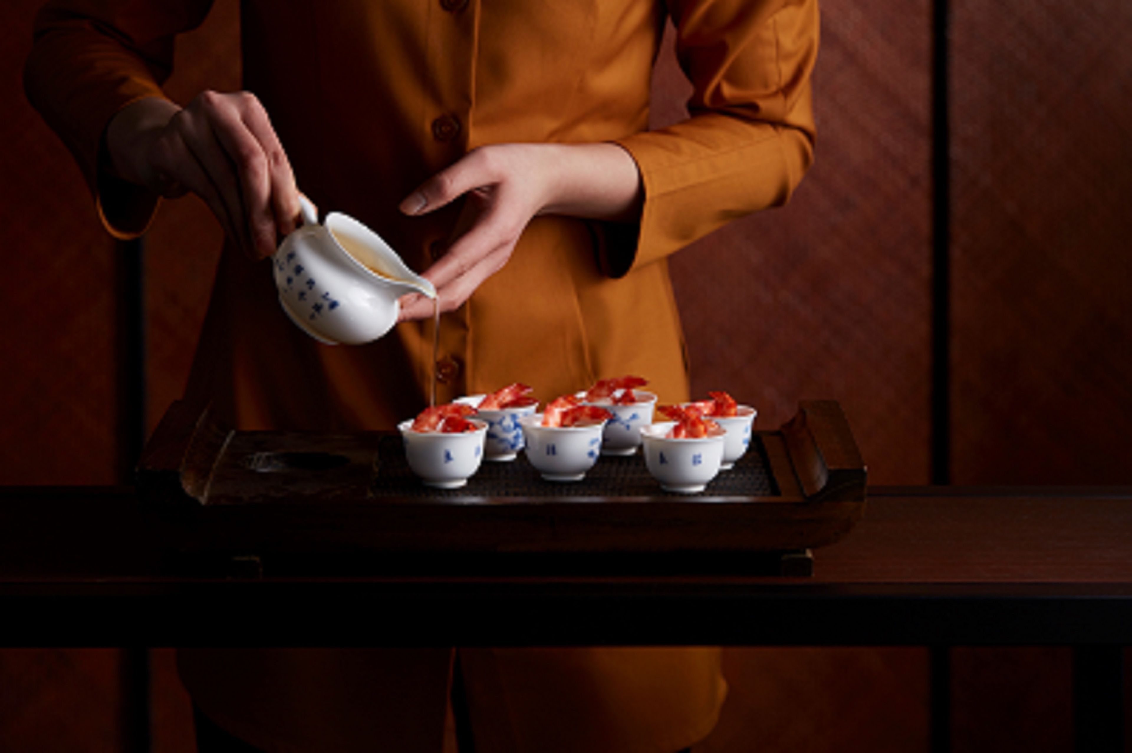 A person in an orange uniform pouring a liquid from a white pitcher into small bowls with blue patterns.