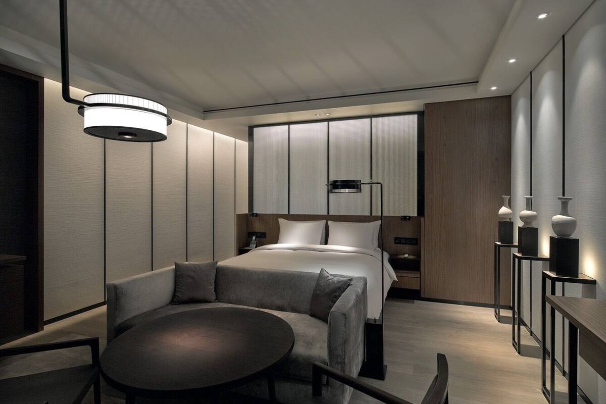 A modern hotel room with a bed, a round table, and a sofa. There is a nightstand with a lamp and a vase on it.