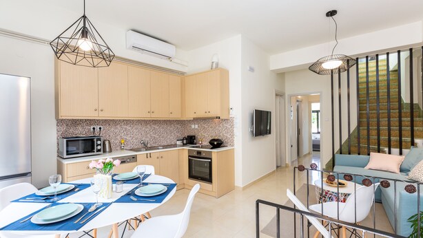 A modern kitchen with wooden cabinets, a dining area with white chairs and blue placemats, and a staircase leading to a living area with a blue sofa.