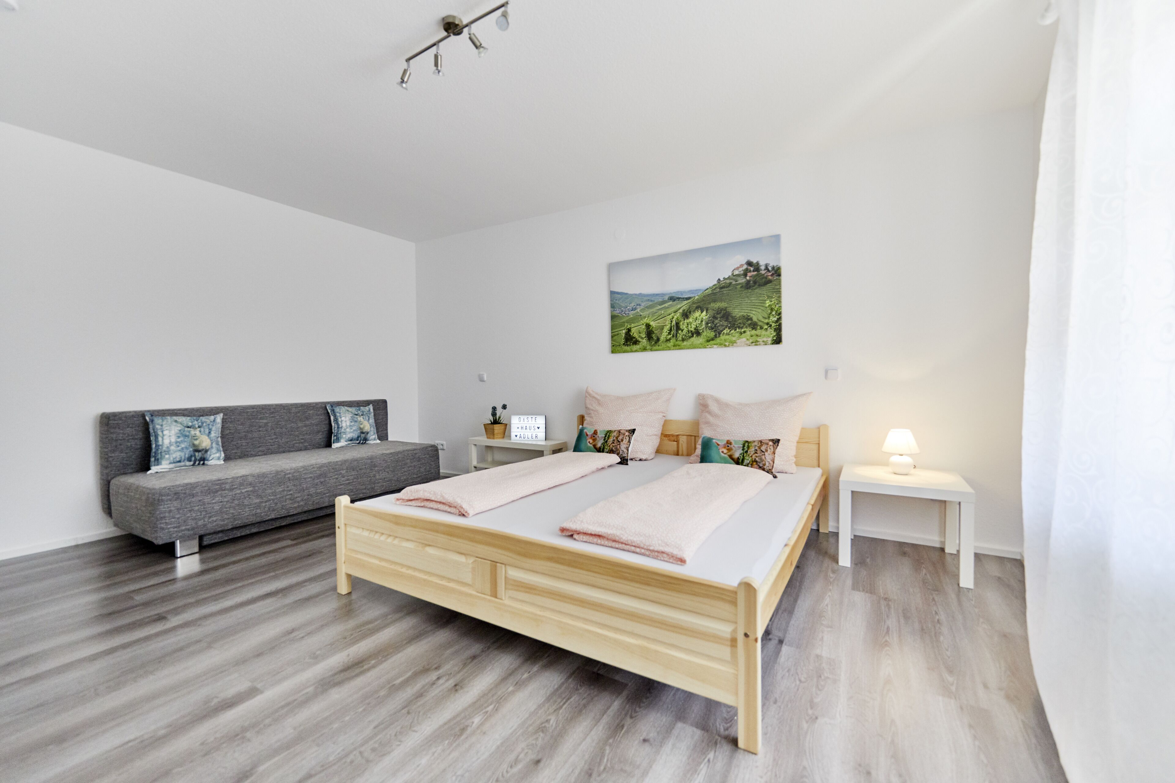 A modern bedroom with a wooden bed, grey sofa, and a landscape painting on the wall.