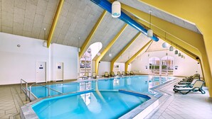 Una piscina interior con techo alto y vigas amarillas, rodeada de sillones de descanso y suelo de baldosas.
