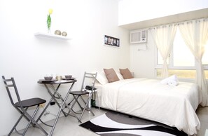 Quarto bem arrumado com cama, uma mesinha com cadeiras, uma estante com objetos decorativos e uma janela com cortinas.