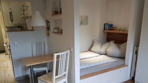 Un espace de vie compact comprenant une table à manger en bois, une chaise blanche, un banc intégré agrémenté de coussins à motifs et une étagère avec des livres et des objets décoratifs.