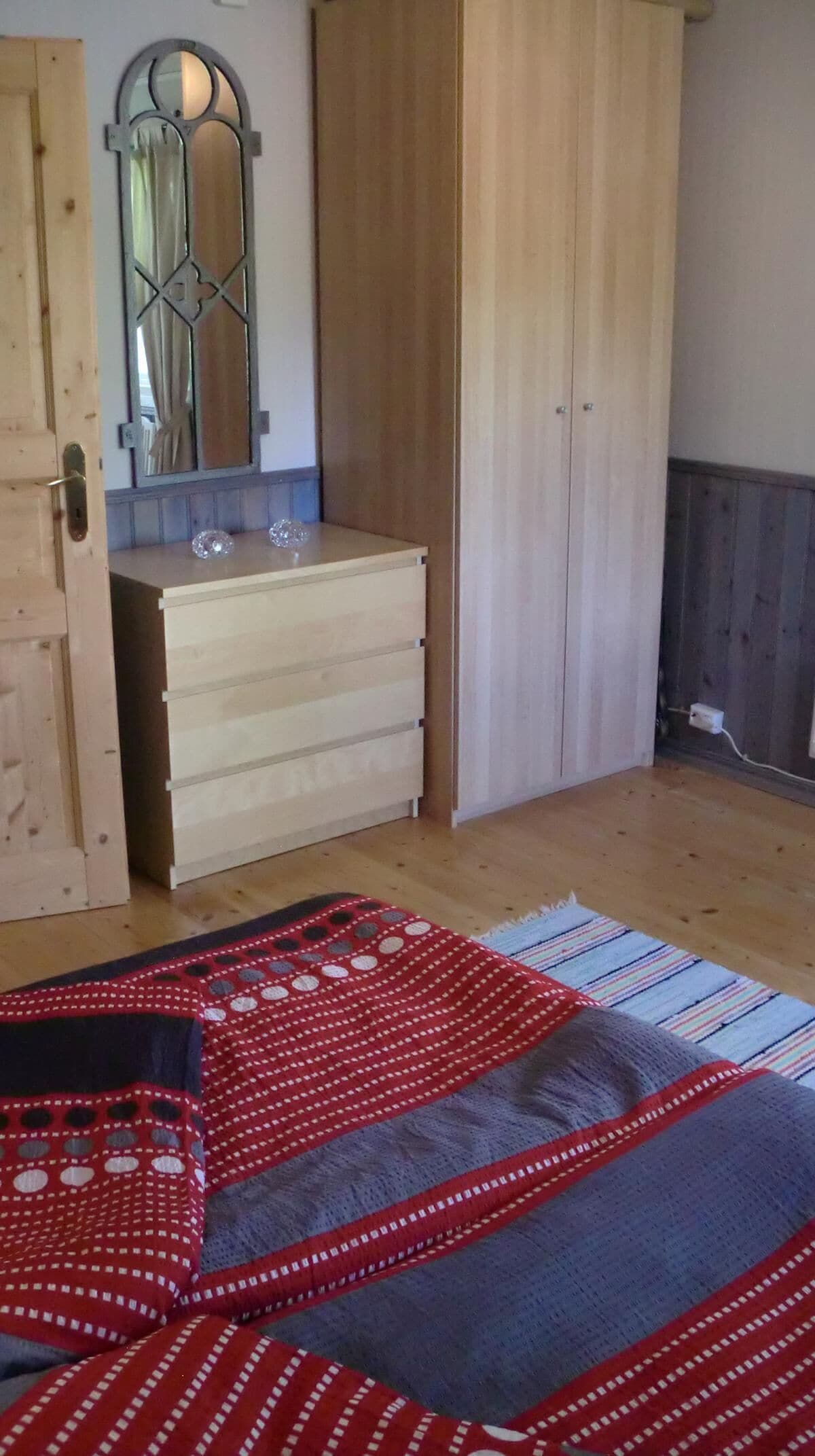 Ein Schlafzimmer mit Holzschrank, Spiegel und Bett mit rot-grauem Musterdecken.