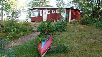 Une cabane en bois rouge, avec une porte blanche, entourée de verdure et à proximité d’un canoë.