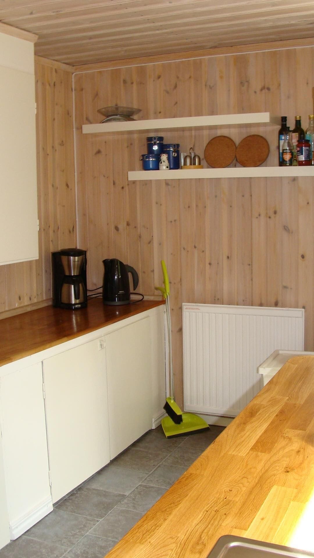 Una cocina con armarios de madera, cafetera, hervidor, escoba y estantes con utensilios de cocina.