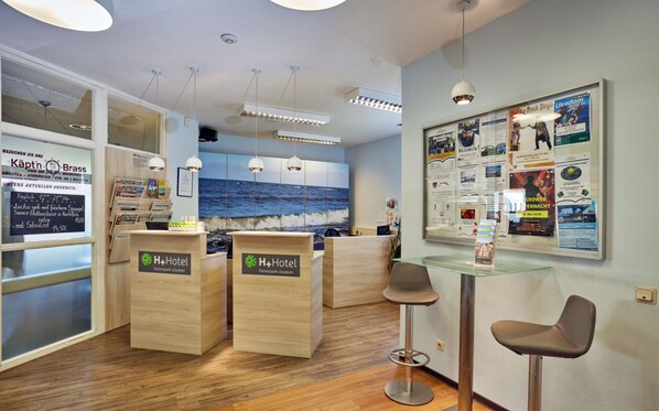 A hotel reception area with wooden reception desks, a glass partition, and a wall with various informational posters.