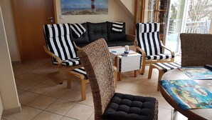 Un espacio de sala cómodo con sillas a rayas y un sofá, una pequeña mesa con un juego de mesa, y un ventanal con vista a una playa y un faro.