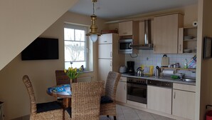 Una cocina con comedor, que incluye una mesa con sillas, un televisor montado en la pared, un ventanal con vistas a árboles y un ventilador de techo.