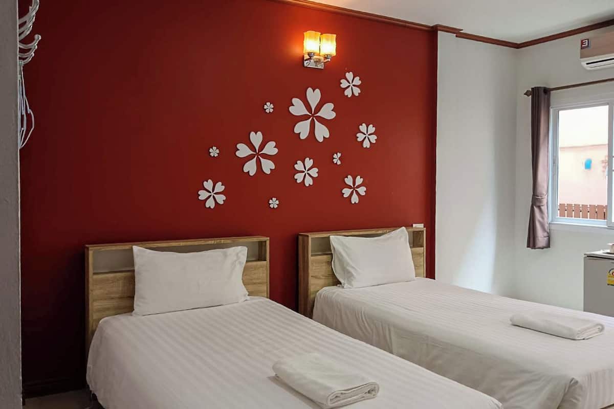 Une chambre d’hôtel avec deux lits, un mur rouge orné de motifs floraux blancs, une fenêtre avec des rideaux et un luminaire fixé au mur.