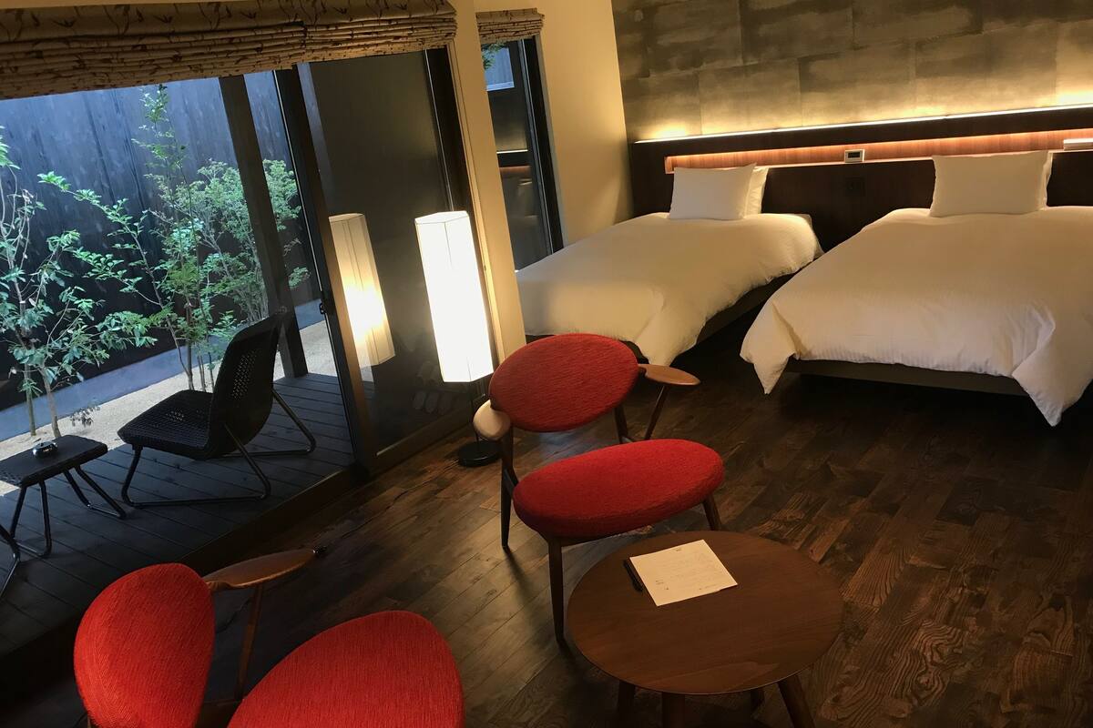 A hotel room with two beds, a wooden floor, a red chair, a small table, and a view of greenery outside.