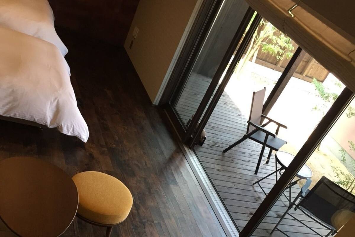 A room with a wooden floor, a yellow armchair with cushions, a small round table, and a bed with white bedding.