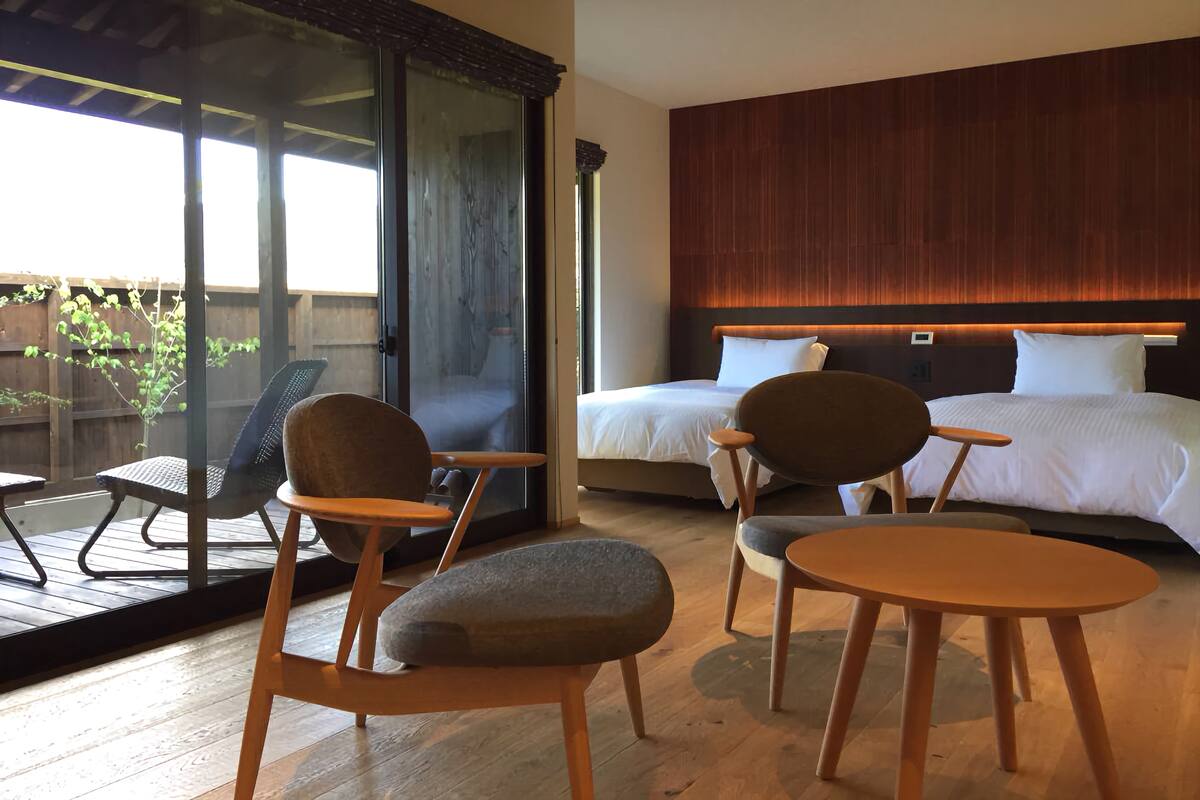 A hotel room with a large bed, two chairs, a small table, and a sliding glass door leading to an outdoor area.