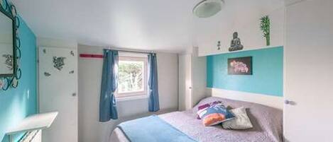 A bedroom with a bed, a window with curtains, a mirror, and a wall-mounted shelf with decorative items.